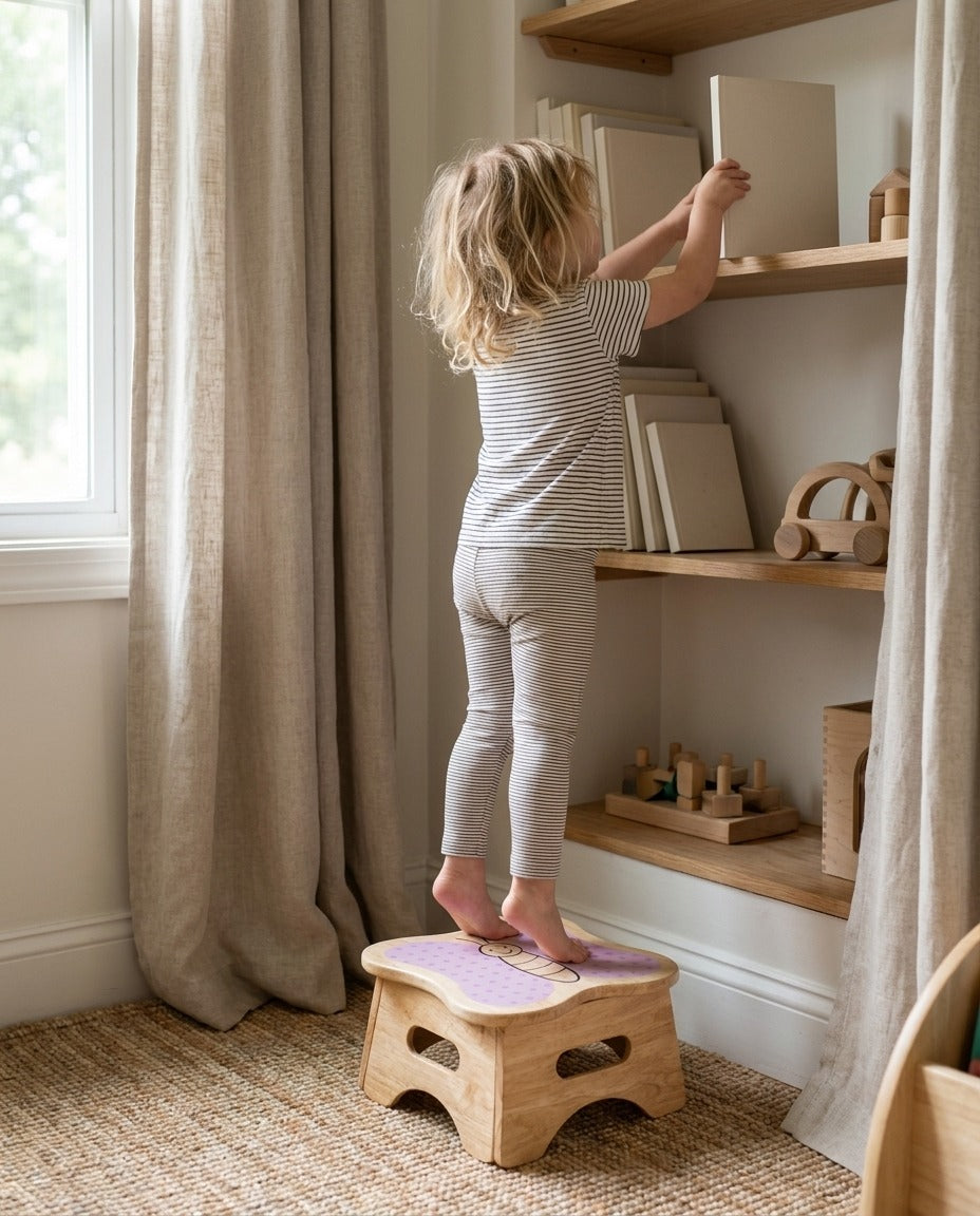 A young girl stands on the Mei butterfly step stool to reach a picture book on a high shelf.