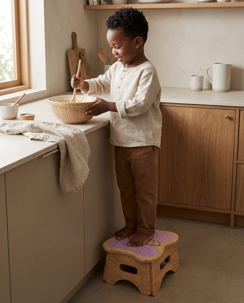A young boy stands on the Mei butterfly step stool at the kitchen counter, stirring a mixing bowl alongside a parent.